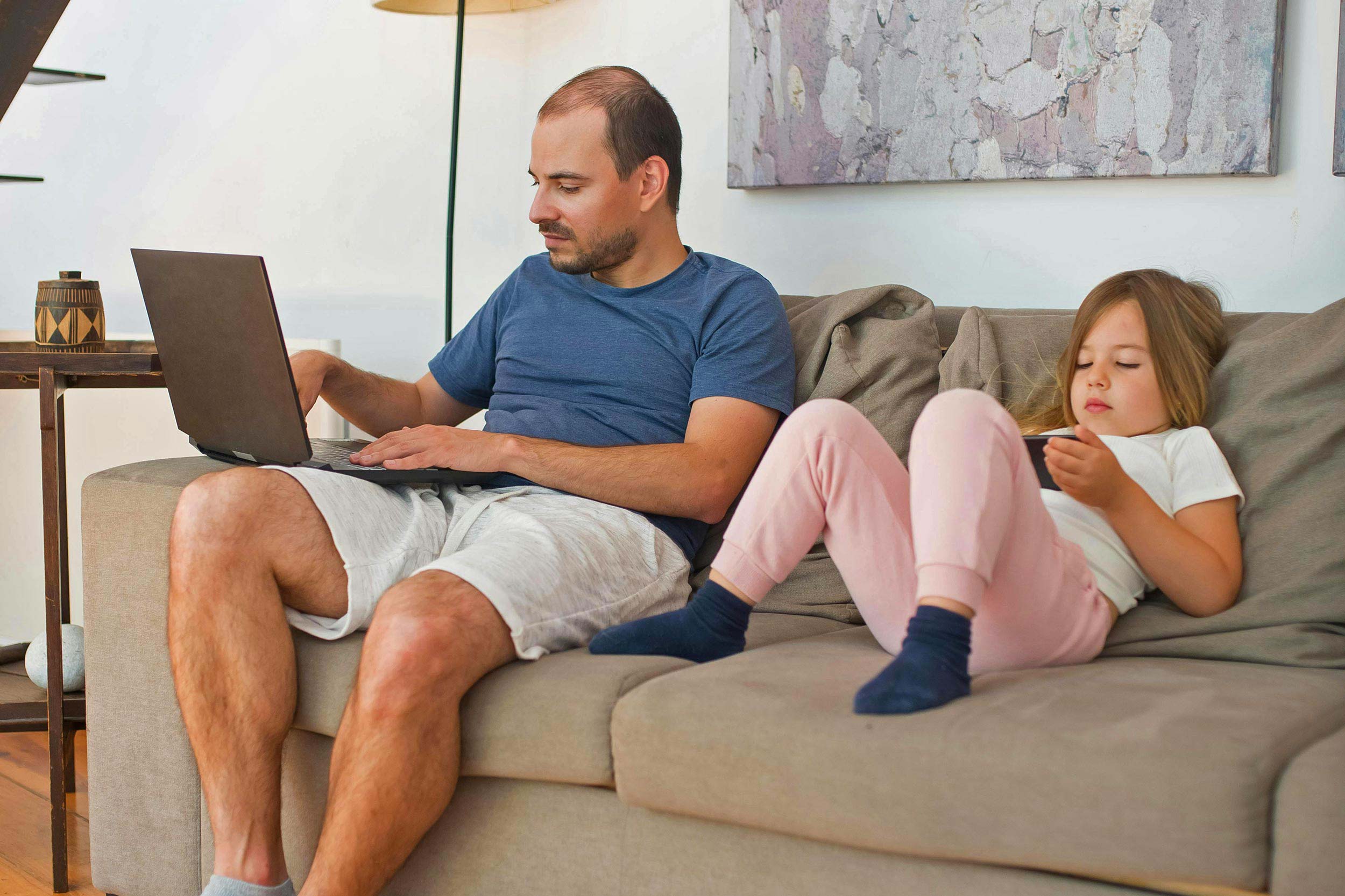 Father using laptop on sofa with child nearby